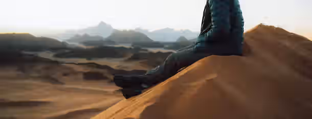 Conquering New Horizons A solitary figure in a jacket sits on a sand dune, gazing at distant mountains, symbolizing an emerging leader's journey to self-improvement.